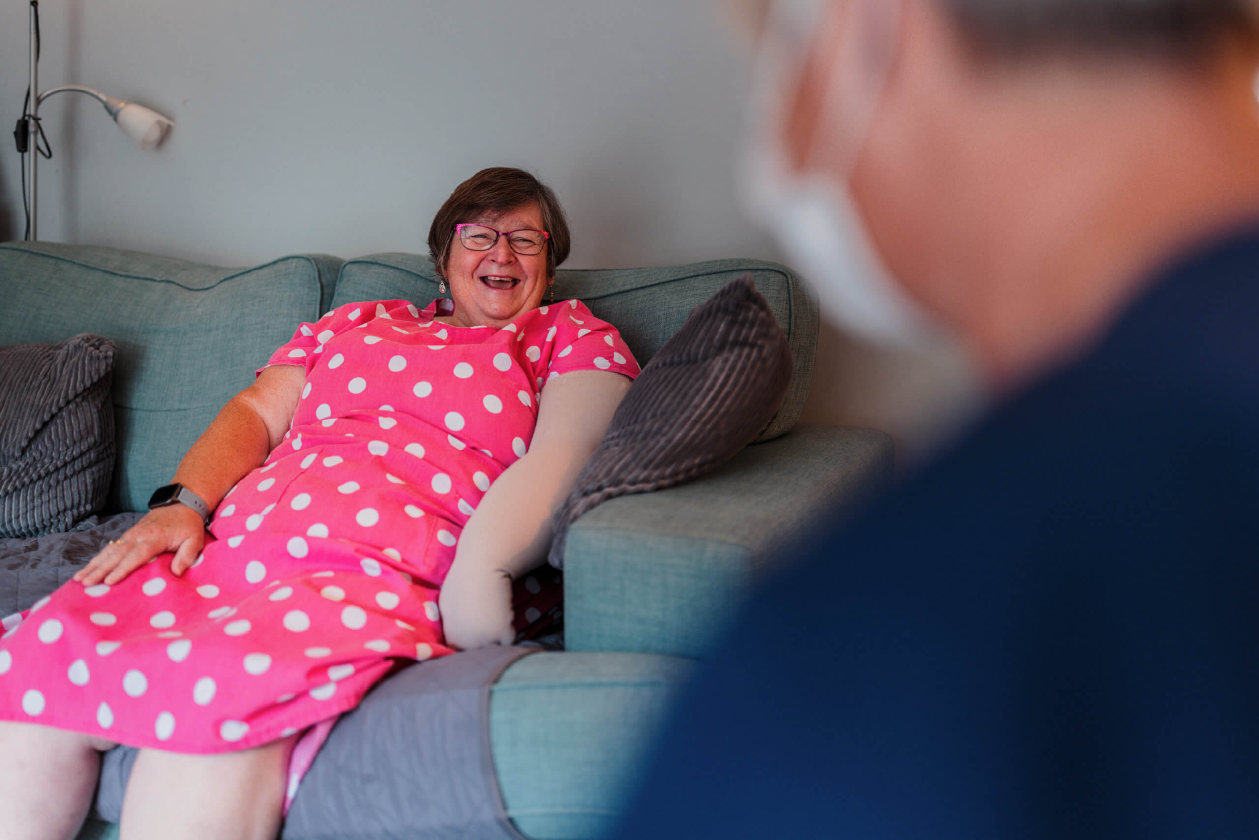 Lady sat on the sofa at home smiling with a nurse in the foreground