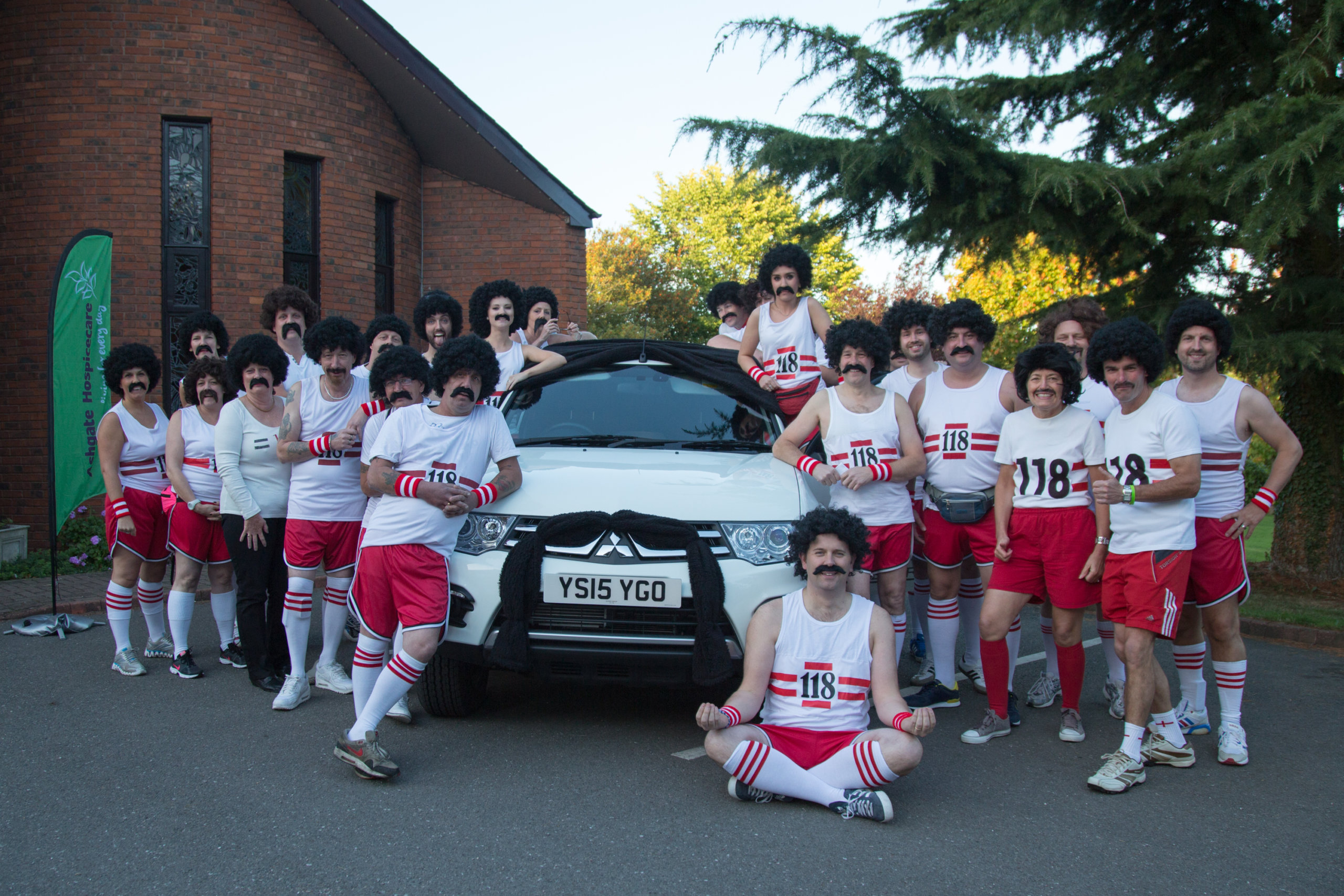 Group of people surrounding a car in fancy dress with wigs and fake moustaches