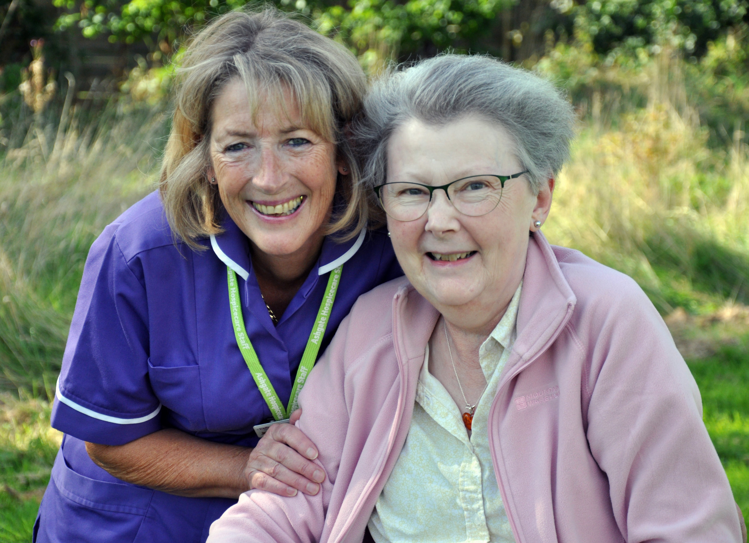 Ashgate Hospice support worker crouched down next to patient who is also smiling. Support worker has her hand on the patients arm