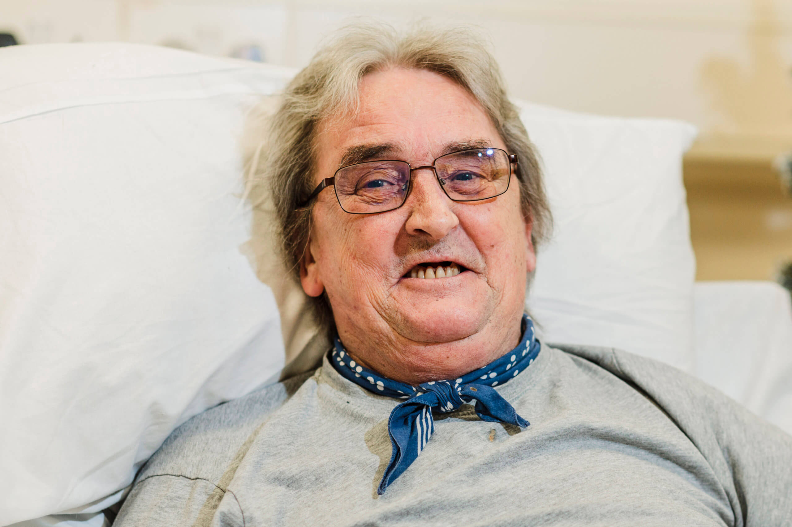 Male patient with glasses and a neckerchief lying in bed smiling