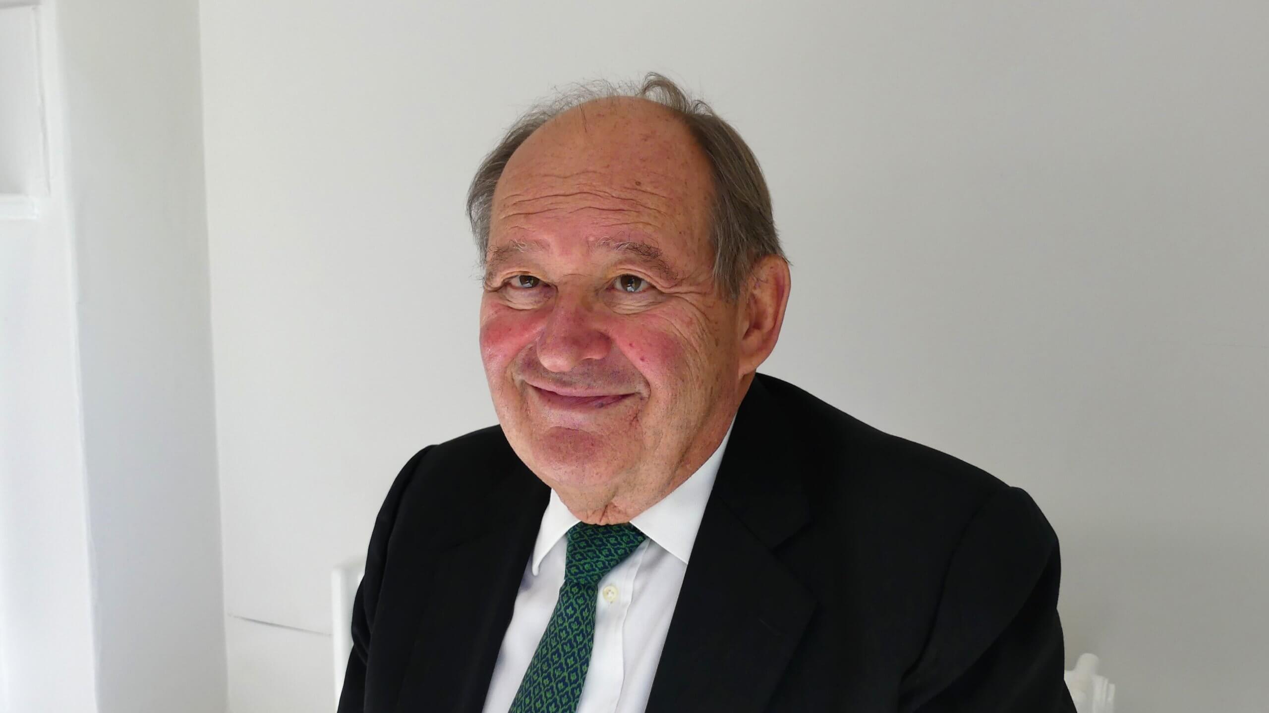 Man in a black suit, white shirt and green tie smiling
