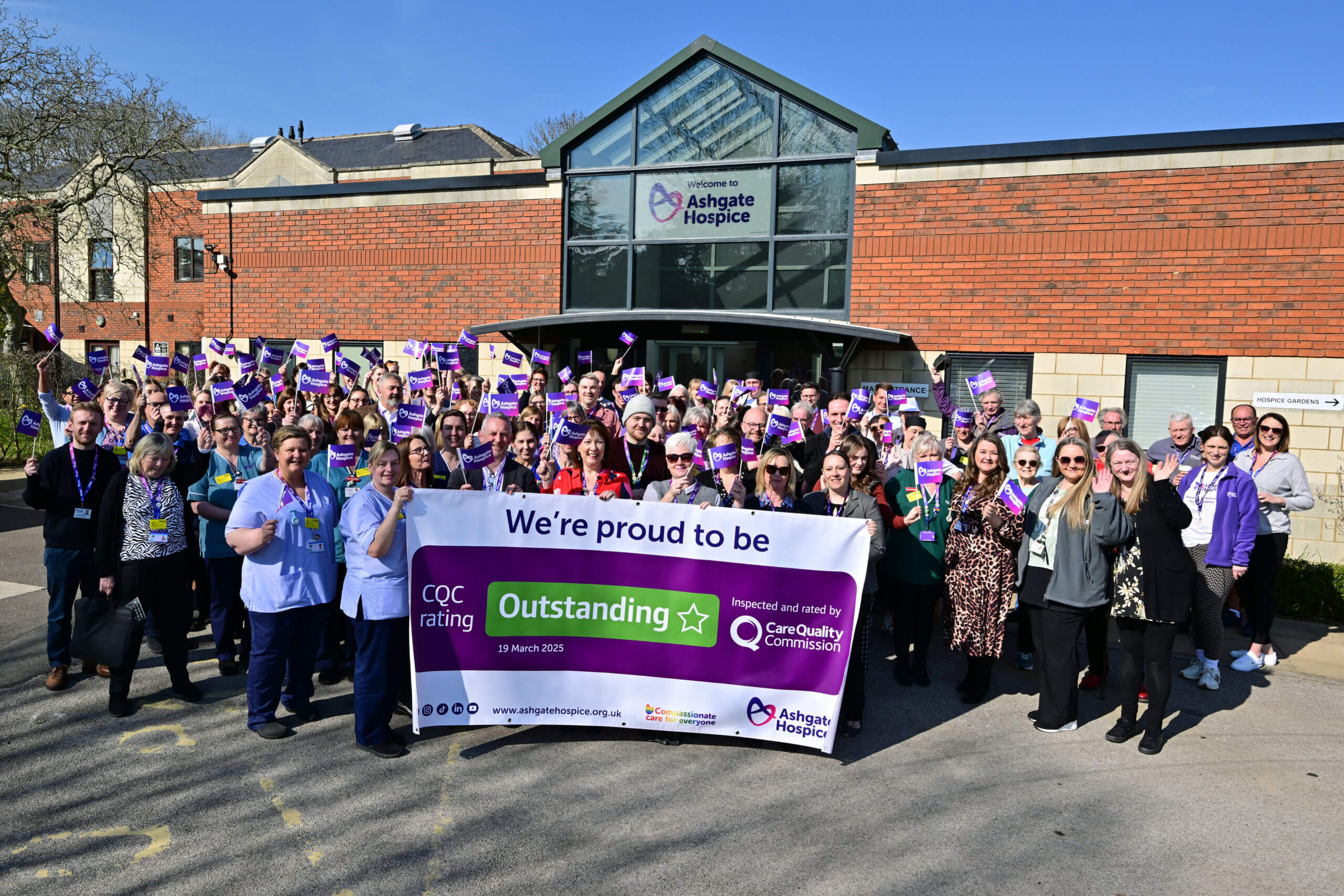 Staff and volunteers outside Ashgate Hospice holding a banner that says: Proud to be outstanding CQC