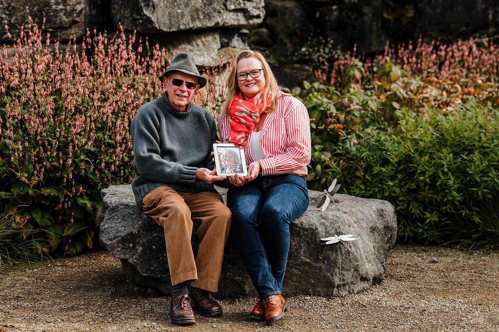 Barry and Clare sitting on a bench at Chatsworth holding a photo of Sue.