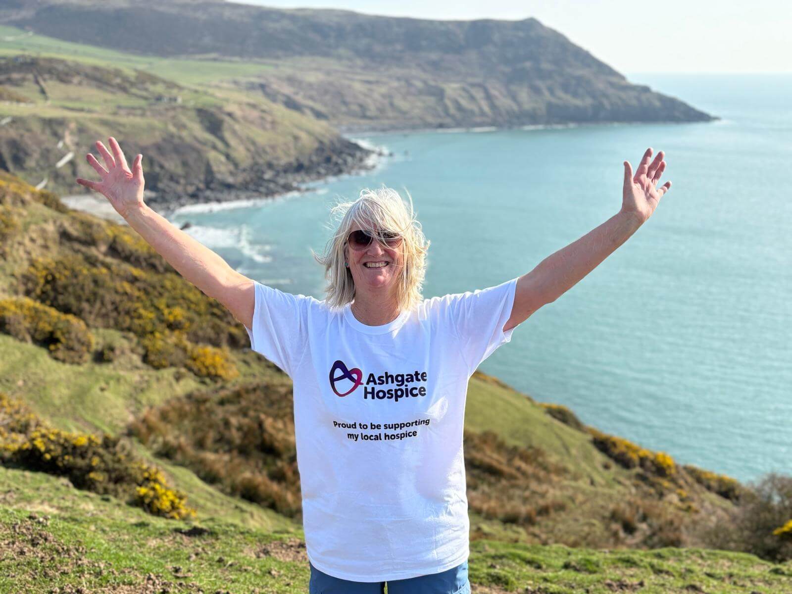Lisa with her arms up in the air, wearing an Ashgate Hospice t-shirt with the coastline in the background.